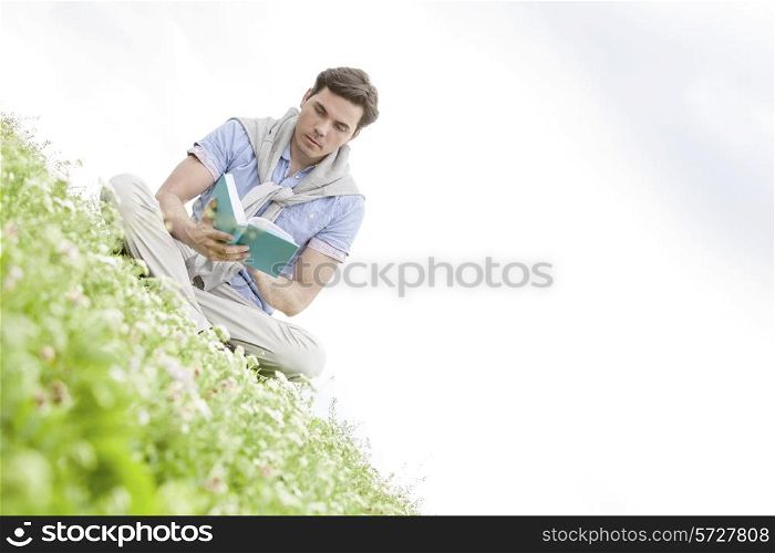Tilt image of young man reading book while sitting on grass against sky