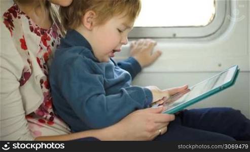 Tilt shot of a little boy sitting on his mothers lap in the train and using tablet PC