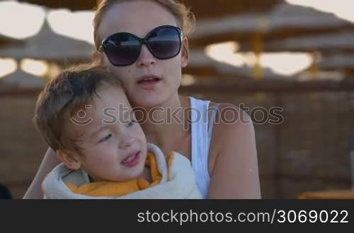 Tilt shot of a lovely family on the beach. Mother and son talking, mom embracing the child wrapped in the towel