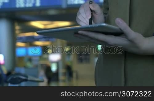 Tilt shot of a woman with tablet computer against flight schedule background. She using pen to type a message