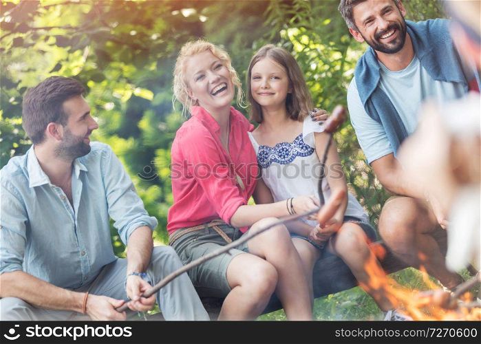 Tilt shot of happy family with male friend roasting sausages over campfire at park