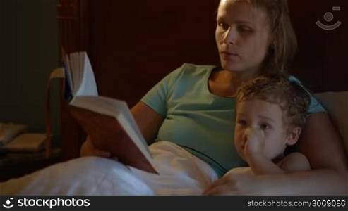 Tilt shot of mother and son in bed at night. Mom reading aloud before bed, boy watching TV