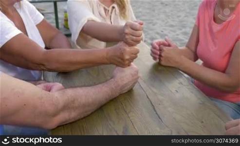 Tilt shot of people sitting at wooden table on the beach and stacking fists in a tower. Teamwork and cooperation