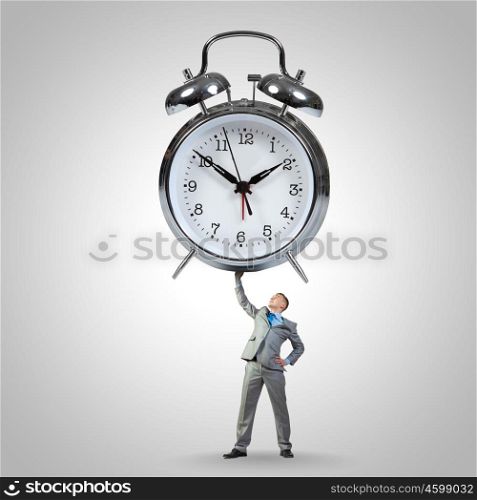 Time concept. Young strong businessman lifting huge alarm clock above head