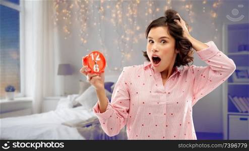time, emotions and bedtime concept - shocked young woman in pajama with alarm clock showing half past six over bed and garland lights at night bedroom background. shocked young woman in pajama with alarm clock