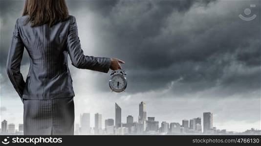 Time for business. Back view of businesswoman holding alarm clock against city background