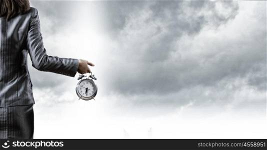 Time for business. Back view of businesswoman holding alarm clock against cloudy background