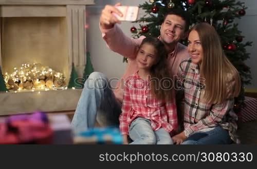 Time for family selfie on Christmas eve. Lovely smiling family sitting on the floor by xmas tree and taking self portrait photo on mobile phone. Adorable daughter and parents making selfie with smartphone as they spend winter holidays together. Dolly