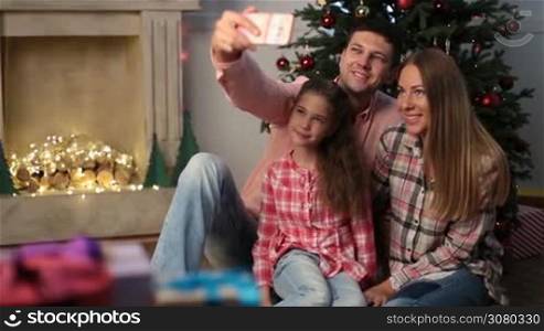 Time for family selfie on Christmas eve. Lovely smiling family sitting on the floor by xmas tree and taking self portrait photo on mobile phone. Adorable daughter and parents making selfie with smartphone as they spend winter holidays together. Dolly