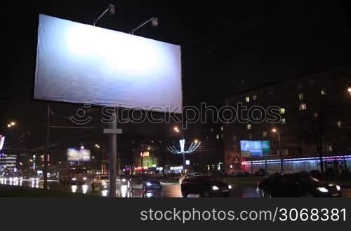 Time lapse of urban scene with an illuminated empty billboard on the side of a street with cars in motion and a block of flats in the background, by night