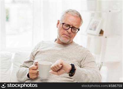 time, morning and people concept - senior man with cup of tea looking at wristwatch at home