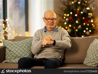time, winter holidays and people concept - senior man looking at wristwatch at home in evening over christmas tree lights on background. senior man looking at wristwatch on christmas