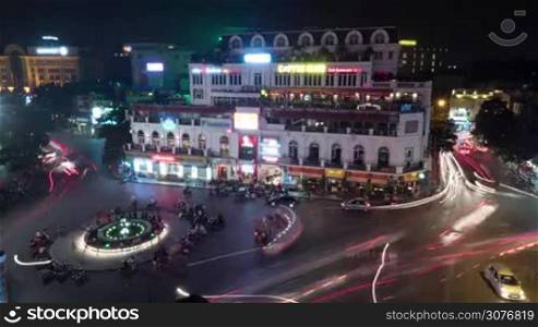 Timelapse high angle shot of road traffic at night. Illuminated fountain with bikes on the city square and transport moving around it. Hanoi, Vietnam