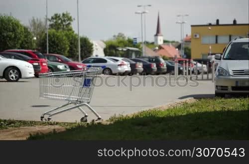 Timelapse of cars and people traffic on parking zone. Empty shopping cart standing in foreground