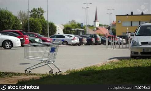 Timelapse of cars and people traffic on parking zone. Empty shopping cart standing in foreground