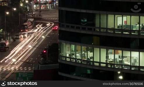 Timelapse shot of night car traffic with modern office business centre in foreground, people working in the office can be seen through the windows. Seoul, South Korea