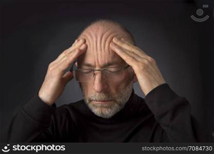 tired - 60 years old man with a beard and glasses massaging his forehead - a headshot against a black background