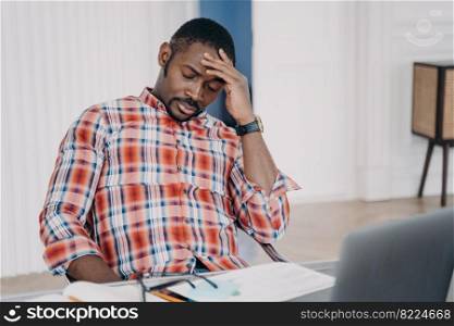 Tired african american businessman suffering from headache, after working at laptop, sitting at workplace. Overworked black male guy touching head, pondering problem. Stress at work concept.. Tired african american male employee suffering from headache after working at laptop. Stress at work