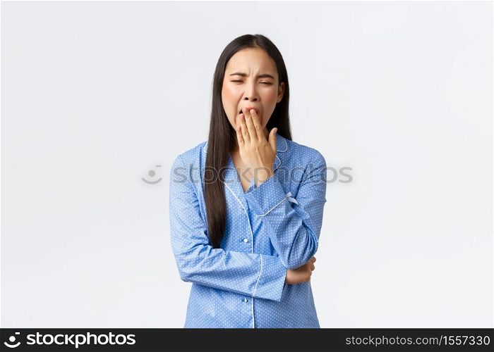 Tired and bored cute asian girl in blue pajamas yawning from boring stories at sleepover party, got exhausted, showing lack of interest, standing over white background wanna go sleep.. Tired and bored cute asian girl in blue pajamas yawning from boring stories at sleepover party, got exhausted, showing lack of interest, standing over white background wanna go sleep
