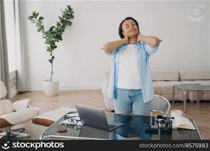 Tired but satisfied freelancer is relaxing after work with her eyes closed. Young european woman is working at home office. Business lady gets up from desk, massaging neck and smiling.. Freelancer is relaxing after work with her eyes closed. Business lady massaging neck and smiling.