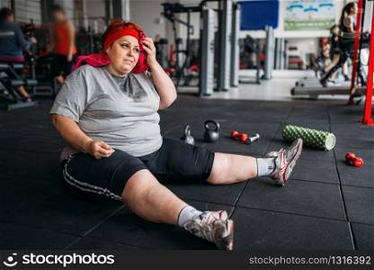 Tired fat woman sits on the floor in gym. Calories burning, obese female person in sport club