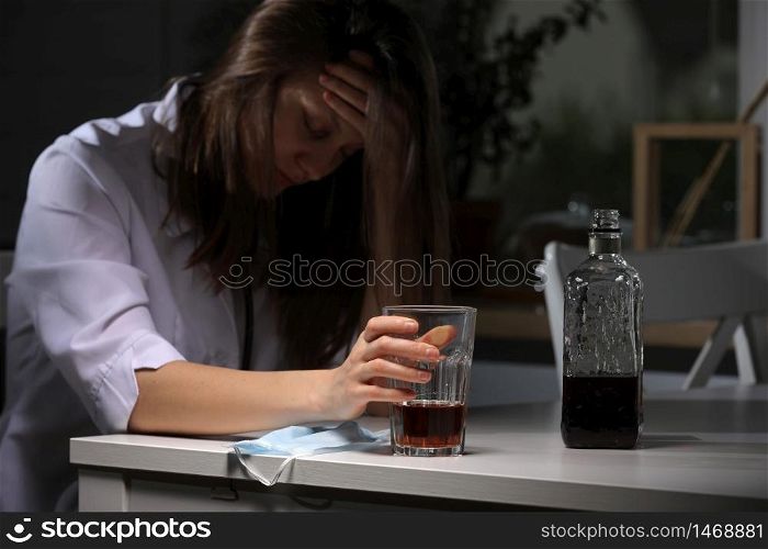 tired female doctor sitting at the table and hold glass with whiskey or cognac at home after hard work, depressed female drinking strong alcohol suffering from coronavirus problems. tired female doctor sitting at the table and hold glass with whiskey or cognac at home after hard work, depressed female drinking strong alcohol suffering from coronavirus problems.