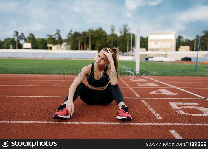 Tired female jogger sitting on the ground, training on stadium. Woman doing stretching exercise before running on outdoor arena