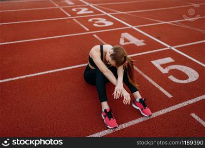 Tired female jogger sitting on the ground, training on stadium. Woman doing stretching exercise before running on outdoor arena. Tired female jogger sitting on the ground, stadium