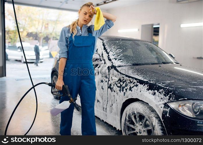 Tired female washer in uniform with foam gun in hands, car wash. Woman washes vehicle, carwash station, car-wash business