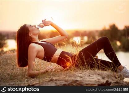Tired girl athlete drinks water from a bottle while lying on the lake at sunset. Tired girl athlete drinks water from bottle