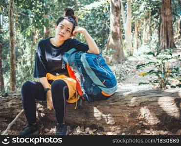 Tired hiking woman resting on log wood in forest with large backpack.