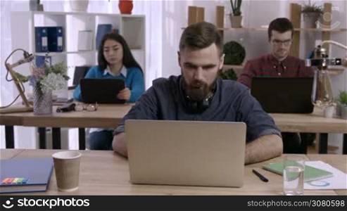 Tired hipster businessman with beard stretching arms in front of laptop while sitting at the desk in modern office. Freelancer stretching and relaxing in office as his colleagues working on background