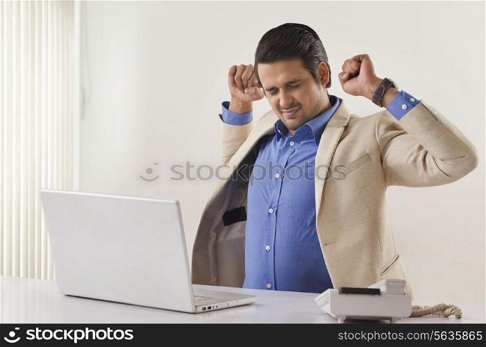 Tired Indian businessman stretching with laptop at office desk