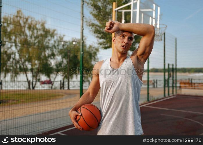 Tired male basketball player with ball on outdoor court, front view. Male athlete in sportswear on streetball training, summer stadium