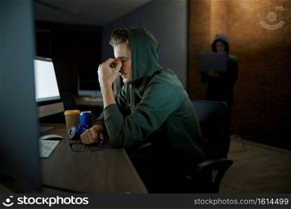 Tired male internet hacker in hood sitting at monitors. Illegal web programmer at workplace, criminal occupation. Data hacking, cyber security. Tired male hacker in hood sitting at monitors