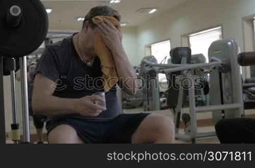 Tired man having a break after workout in a gym. He wiping the head with a towel and drinking water. Another man preparing for exercise