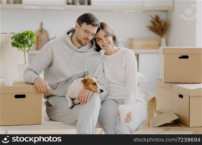 Tired married family couple drink aromatic takeaway coffee, sit closely to each other, pose with pedigree dog near big carton boxes, pose against kitchen interior, move into new modern apartment