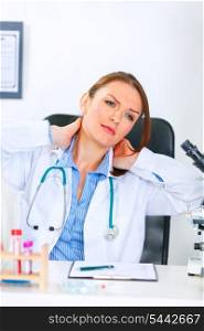 Tired medical doctor woman sitting at office table and massaging her neck&#xA;