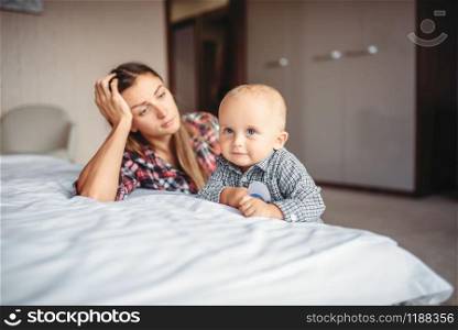 Tired mother looks on her little child playing on the bed, motherhood problems. Depressed mom and son together at home, parenthood trouble