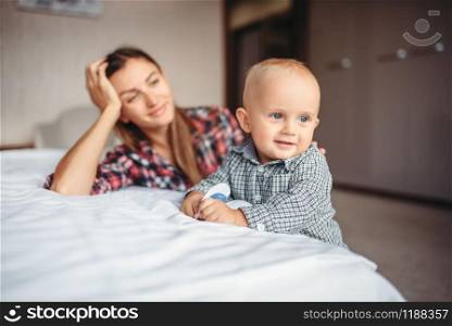 Tired mother looks on her little child playing on the bed, motherhood problems. Depressed mom and son together at home, parenthood trouble. Tired mother looks on her child playing on the bed