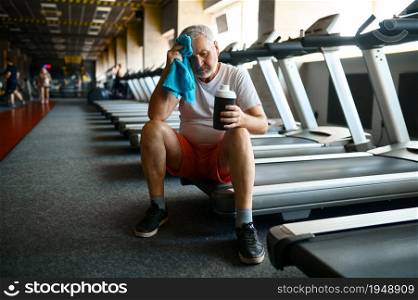 Tired old man with towel drinks water after treadmill, gym interior on background. Sportive grandpa on fitness training in sport center. Healty lifestyle, health care, elderly sportsman. Tired old man with towel drinks water in gym