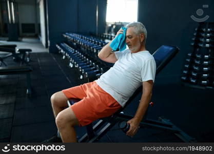 Tired old man with towel leisures after workout, gym interior on background. Sportive grandpa on fitness training in sport center. Healty lifestyle, health care, elderly sportsman. Tired old man with towel leisures after workout