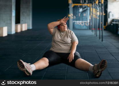 Tired overweight woman sits on the floor in gym. Calories burning, obese female person in sport club, fat people. Tired overweight woman sits on the floor in gym