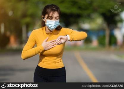 tired runner woman looking smart watch and wearing medical mask to protect Coronavirus Covid-19  pandemic