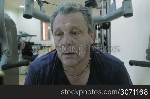 Tired sweaty man having a break and catching breath between sets on exercise machinne. Young man doing situps in background