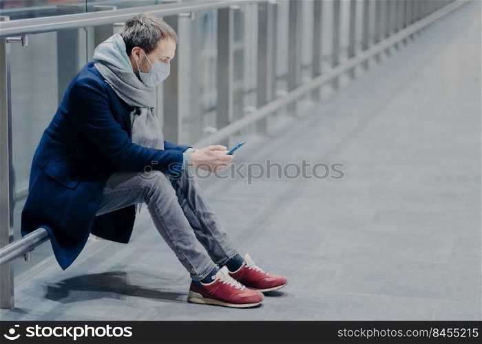 Tired tourist takes break, uses mobile phone, dressed in jacket, jeans and sneakers, wears protective mask against coronavirus, cares about health in public place, reads information. Safety concept