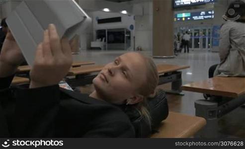 Tired woman lying on the bench in airport lounge and using tablet computer while waiting for the flight