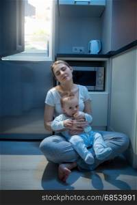 Tired woman sitting on floor at kitchen at night and feeding her baby