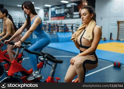Tired woman with towel on a stationary bike in gym. People on fitness workout in sport club, athletic girls in sportswear on training indoors. Tired woman with towel on a stationary bike in gym