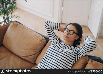 Tired young female relaxing on cozy sofa in living room at home, holding hands behind head. Woman wearing glasses pauses, takes a break during household chores, resting on comfortable couch.. Tired female rests on sofa, takes a break, enjoys recreation, sitting on comfortable couch at home